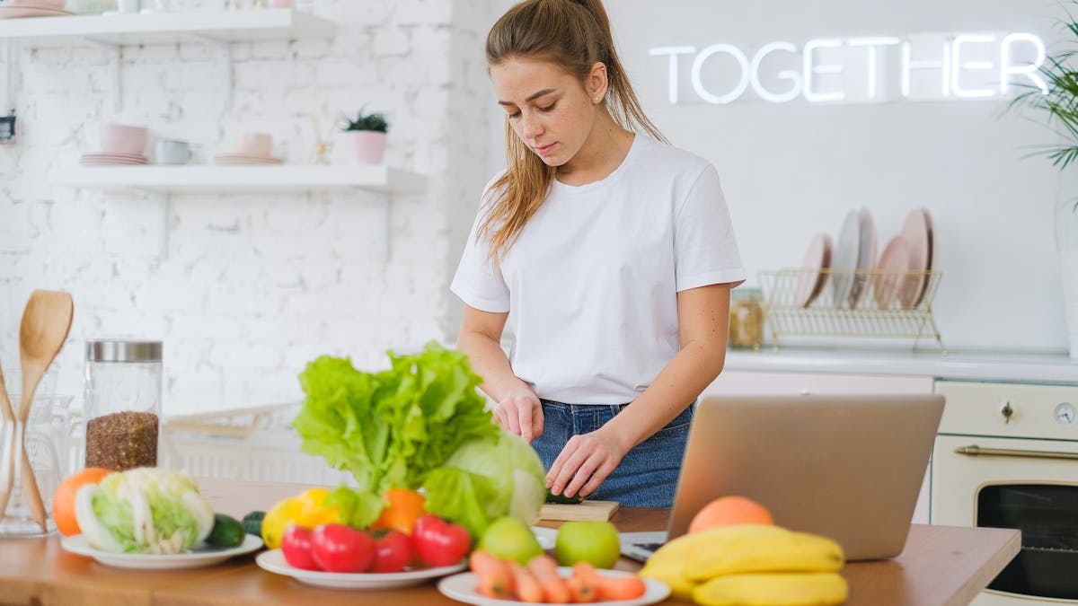 Woman in white t-shirt preparing fresh vegetables in a bright kitchen with a laptop.